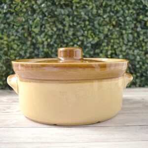 May include: A tan and brown ceramic casserole dish with a lid. The dish has two small handles on either side and a rounded shape. The lid has a small, rounded handle. The dish is likely for cooking and serving food.