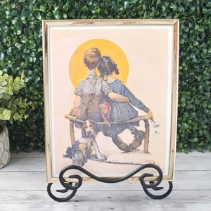 May include: Framed print featuring a boy and girl sitting on a bench with a dog, set against a yellow sun. The print is in a gold-colored frame and displayed on a black metal stand. The artwork has a vintage, nostalgic feel.