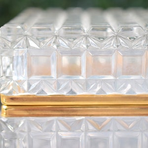 K&ouml;nnte beinhalten: Eine transparente, rechteckige Glasbox mit einem geometrischen, strukturierten Muster. Die Box hat ein goldfarbenes Band in der Mitte. Deckel und Boden sind mit einem sich wiederholenden Quadrat- und Rautenmuster verziert.