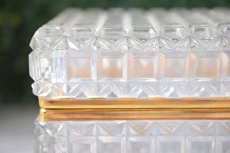 K&ouml;nnte beinhalten: Eine transparente, rechteckige Glasbox mit einem geometrischen, strukturierten Muster. Die Box hat ein goldfarbenes Band in der Mitte. Deckel und Boden sind mit einem sich wiederholenden Quadrat- und Rautenmuster verziert.