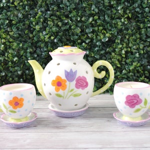 May include: A ceramic tea set with a teapot and two teacups, each decorated with colorful floral designs. The teapot and cups are white with green, purple, and orange accents. The set is designed for decorative use.