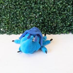 May include: A plush toy shaped like a beetle with a bright blue body and a darker blue shell. The insect features six legs with black tips, two antennae, and a single visible eye. The soft fabric construction gives the creature a rounded form.