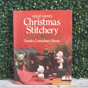 May include: A red book titled "Scrap Saver's Christmas Stitchery" by Sandra Lounsbury Foose. The cover features a sewing machine, teddy bears, and spools of thread. The background is a green hedge.