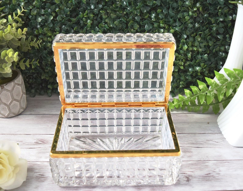 K&ouml;nnte beinhalten: Eine rechteckige, transparente Glasschmuckschatulle mit goldfarbenem Rand. Die Schatulle hat ein Gittermuster an den Seiten und am Deckel. Der Innenboden hat ein Sternmuster. Auf einer wei&szlig;en Holzoberfl&auml;che ausgestellt.