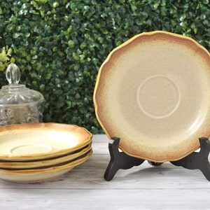 May include: A set of vintage ceramic saucers with a scalloped edge and a speckled beige surface. The rim is a darker brown. One saucer is displayed on a black stand, while three are stacked. The background features greenery.