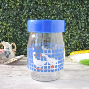 May include: Clear plastic jar with a blue lid. The jar features a blue and white checkered pattern with white geese. The jar is on a white wooden surface with a green hedge background.