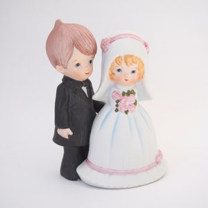May include: A ceramic wedding cake topper featuring a bride and groom. The groom wears a black tuxedo with a bow tie, and the bride is in a white dress with a veil and holds a bouquet of pink roses. The figures have painted facial features.