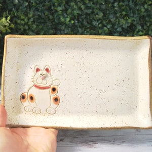 May include: Rectangular ceramic serving tray with a cream-colored speckled surface and a raised, smiling cat design. The cat has orange spots and a red collar. The tray has a light brown rim.