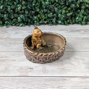 May include: A ceramic dog figurine sits inside a brown, basket-style bowl. The dog is brown and tan, with detailed fur and facial features. The bowl has a woven texture and a patterned interior, creating a rustic aesthetic.