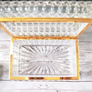 K&ouml;nnte beinhalten: Eine rechteckige, transparente Schmuckschatulle aus Glas mit goldfarbenem Rand, ge&ouml;ffnet. Der Deckel und der Boden weisen ein detailliertes Schliffmuster auf. Der Boden hat ein Sonnenstrahl-Design. Die Schatulle steht auf einer wei&szlig;en Holzoberfl&auml;che.