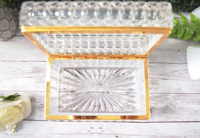 K&ouml;nnte beinhalten: Eine rechteckige, transparente Schmuckschatulle aus Glas mit goldfarbenem Rand, ge&ouml;ffnet. Der Deckel und der Boden weisen ein detailliertes Schliffmuster auf. Der Boden hat ein Sonnenstrahl-Design. Die Schatulle steht auf einer wei&szlig;en Holzoberfl&auml;che.
