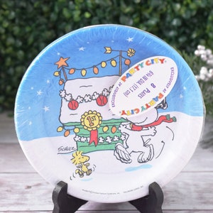 May include: A package of eight round paper plates featuring a winter scene with Snoopy and Woodstock. The plates have a blue border with a snowy landscape design and the text "PARTY CITY". Each plate is 6 3/4 inches in diameter.
