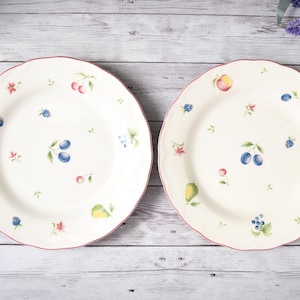 May include: Two white ceramic plates with scalloped edges and a red trim. Each plate is decorated with colorful fruit and floral designs, including plums, cherries, and peaches. The plates are set against a light wood background.