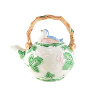 May include: A white ceramic teapot with a bird-shaped lid and a branch-like handle. The teapot is decorated with green leaves and pink flowers. The bird is blue and white.