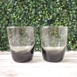May include: Two clear drinking glasses with a dark gray base and a lighter gray gradient. The glasses are cylindrical and sit on a white wooden surface, with a green hedge background.