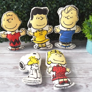 May include: A collection of five fabric character dolls featuring the Peanuts gang. The dolls include Charlie Brown in a red shirt, Lucy in a yellow dress, Linus in a blue striped shirt, Snoopy, and Sally in a red dress. Each doll has a smiling face and outstretched arms.