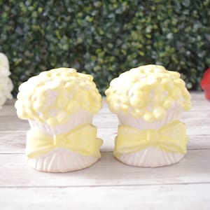 May include: Two white ceramic salt and pepper shakers with a yellow floral design on top and a yellow bow. The shakers are on a white wooden surface with a green hedge background and red apples.