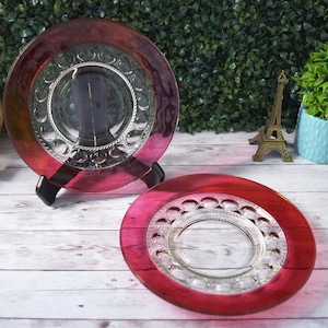 May include: Two decorative glass plates with a red and clear design. The plates are displayed on a wooden surface with a green hedge background. One plate is on a black stand.