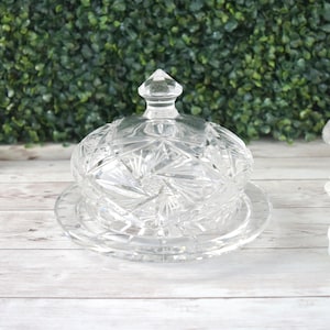 May include: A clear, cut-glass butter dish with a domed lid and a round base. The lid features a pointed finial and a starburst pattern. The dish is designed to hold butter and is a decorative kitchen item.