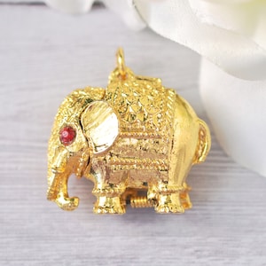 May include: A gold-colored elephant pendant with intricate detailing. The elephant has a red gemstone eye and is adorned with decorative patterns. The pendant is suspended from a small loop, suggesting it's designed to be worn as jewelry.