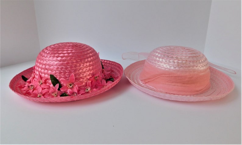 May include: Two pink straw hats with a wide brim. One hat has a pink flower trim and the other has a pink ribbon.