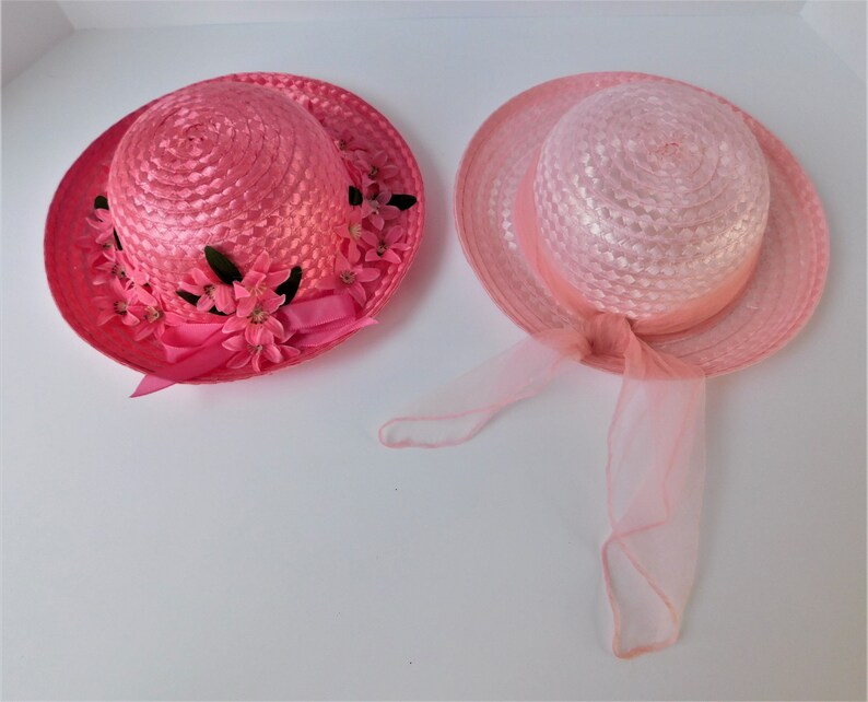 May include: Two pink straw hats with floral embellishments. One hat has a pink ribbon and the other has a pink tulle bow.
