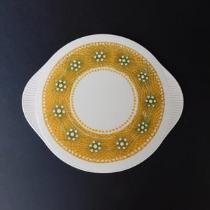 May include: White serving dish with a yellow and green geometric design. The dish has a circular pattern with a white center and two handles. The design features small white dots and a network of yellow lines.