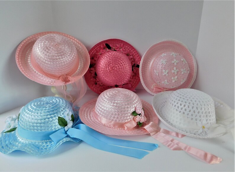 May include: Six straw hats with various colors and embellishments. The hats are pink, white, blue, and a darker pink. Some hats have flowers, ribbons, and other decorative elements.