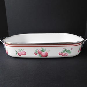 May include: White rectangular baking dish with a red stripe and fruit illustrations. The dish has silver handles and a silver rim. The fruit illustrations include cherries and apples. The dish is on a black surface.