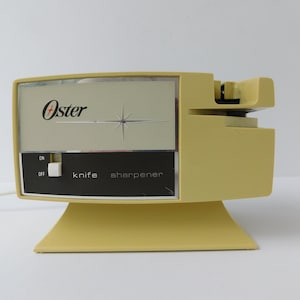 May include: A vintage Oster knife sharpener in a pale yellow color. The appliance has a rectangular face with the Oster logo and a starburst design. The control panel reads "ON", "OFF", and "knife sharpener".