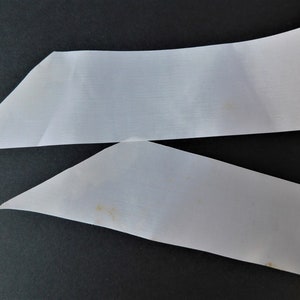 May include: Two white satin ribbon pieces, one folded in a triangle shape, the other straight, with a slight yellow stain on the bottom piece.