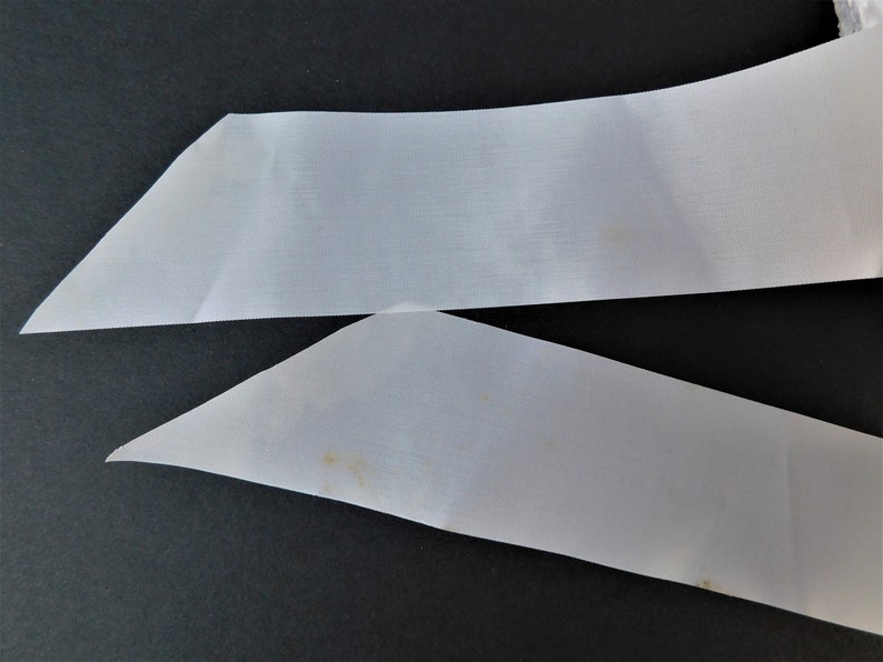 May include: Two white satin ribbon pieces, one folded in a triangle shape, the other straight, with a slight yellow stain on the bottom piece.