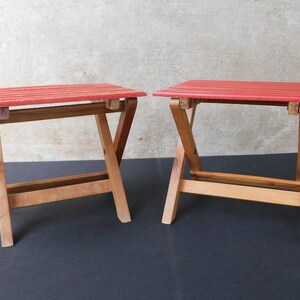 May include: Two red wooden folding stools with a simple design. The stools are made of natural wood and have a red painted top.