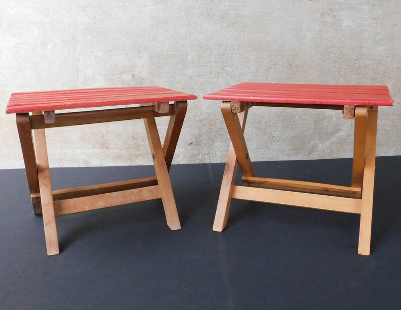 May include: Two red wooden folding stools with a simple design. The stools are made of natural wood and have a red painted top.