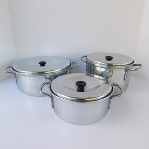May include: Set of three silver pots with lids. The pots are made of metal and have black handles. The pots are nested inside each other.