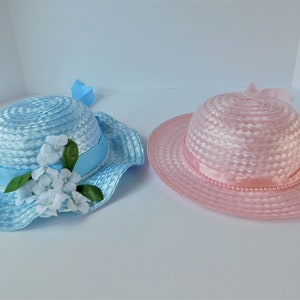 May include: Two straw hats, one blue with white flowers and a blue ribbon, the other pink with a pink ribbon.