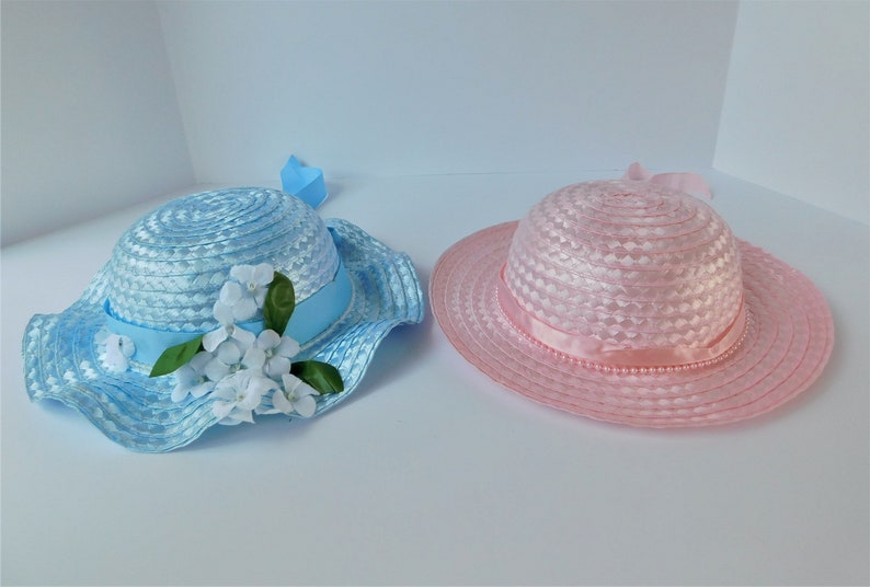 May include: Two straw hats, one blue with white flowers and a blue ribbon, the other pink with a pink ribbon.