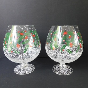 May include: Two clear brandy snifters with a festive Christmas design. The glasses feature green holly leaves and red berries, with a white, snowy effect around the base. The stem and base are clear.