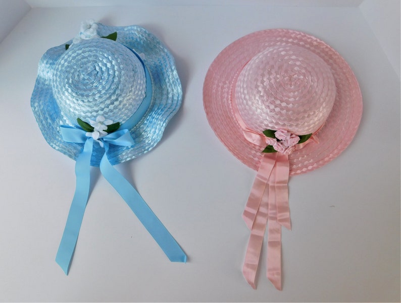 May include: Two straw hats with ribbon ties and flower embellishments. One hat is light blue and the other is pink.