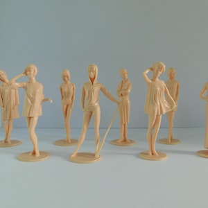 May include: A collection of nine vintage plastic fashion figures in various poses. The figures are a light peach color and stand on small, round bases. They are dressed in different styles, including dresses, coats, and athletic wear.