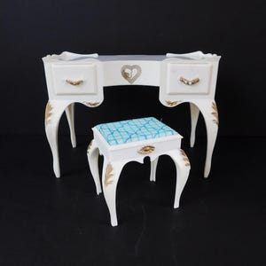 May include: A white vanity desk and matching stool set. The desk has two drawers with gold-colored handles and decorative gold accents. The stool has a blue and white patterned cushion. The set is designed with curved legs and a heart-shaped detail.