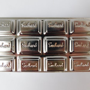 May include: A silver-colored, rectangular mold with 12 individual sections, each embossed with the word "Suchard". The mold is designed to resemble a chocolate bar, with a shiny, metallic finish.