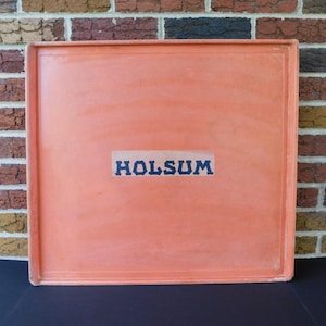 May include: An orange square tray with the word "HOLSUM" in blue block letters. The tray has a slightly raised edge and is set against a brick wall background.