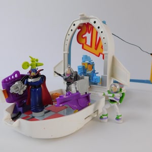 May include: A white toy spaceship playset with several action figures. The figures include Buzz Lightyear, Zurg, and other characters. The playset has a red and yellow sign that says "24/7". The playset is made of plastic.