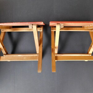 May include: Two wooden folding stools with red painted seats. The stools are made of light brown wood and have a simple, functional design.