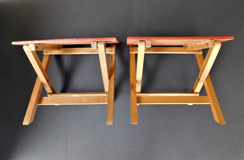 May include: Two wooden folding stools with red painted seats. The stools are made of light brown wood and have a simple, functional design.