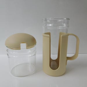 May include: Two clear glass containers with beige lids and holders. One is a small jar with a rounded lid. The other is a tall glass in a beige holder with a handle. The items are on a white surface.