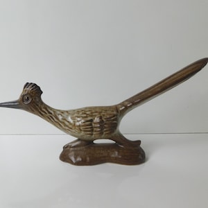 May include: A ceramic roadrunner figurine featuring a glossy brown and tan glaze. The bird is depicted in a horizontal stance with a long, pointed tail, a crest on its head, and a sharp beak, all mounted on a solid, dark brown base.