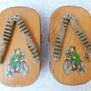 May include: A pair of wooden sandals with black and white striped fabric straps. Each sandal features a cartoon image of a child riding a bicycle.