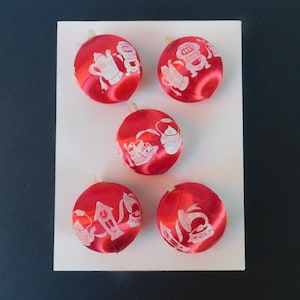 May include: Five round red satin ornaments arranged on a white rectangular card. Each ornament features white printed illustrations of household items including a coffee pot, a stove, a kettle, a coffee grinder, a grandfather clock, and a gramophone.